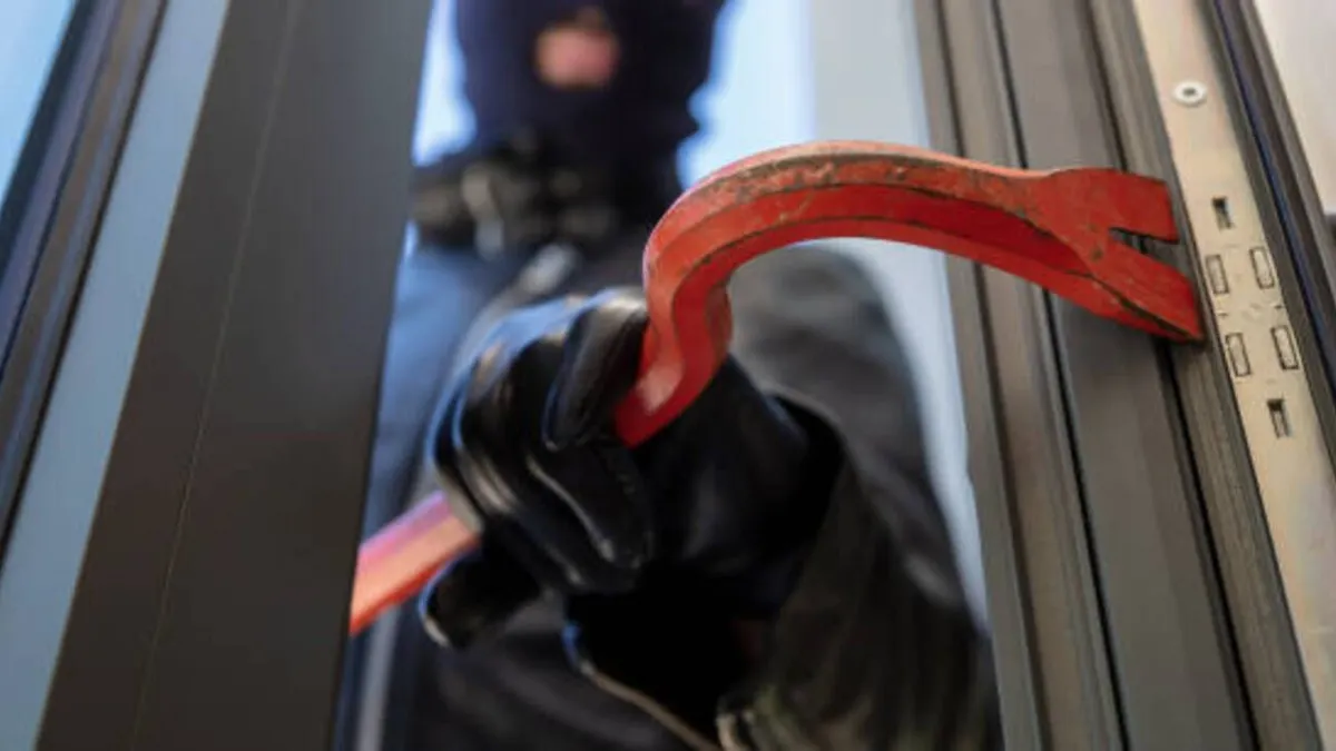 Burglary with crowbar. Burglar in balaclava breaking a glass door. Masked housebreaker entering a house for stealing in the daytime.