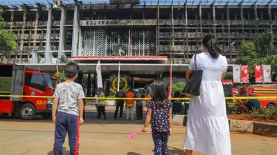 Seorang ibu membawa anak-anaknya untuk melihat gedung utama Kejaksaan Agung RI yang habis terbakar di Jalan Sultan Hasanuddin, Jakarta Selatan, Minggu (23/8). [Suara.com/Alfian Winanto]